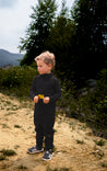 Enfant portant un ensemble thermique évolutif à col roulé en laine de mérinos noire Timininous, debout dans un environnement extérieur forestier. Cet ensemble offre une protection optimale contre le froid grâce à son col roulé, idéal pour les aventures en plein air en toutes saisons."