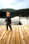 Enfant portant la combinaison thermique évolutive en laine de mérinos noire Timininous, debout sur un quai en bois au bord d'un lac entouré de montagnes. Parfaite pour toutes les saisons, cette combinaison offre confort et adaptabilité grâce à sa conception en mérinos doux, idéal pour les aventures extérieures.